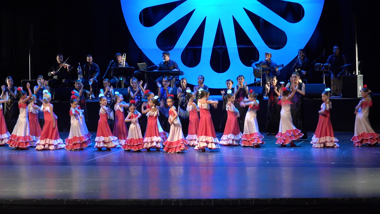 Girls in pink dresses performing on a lit stage with musicians behind them.