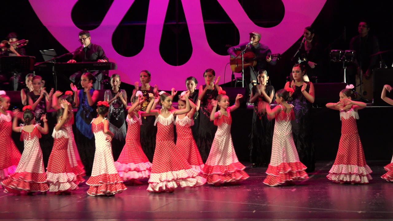 Children in red and white dresses dance on a pink-lit stage with musicians behind them.