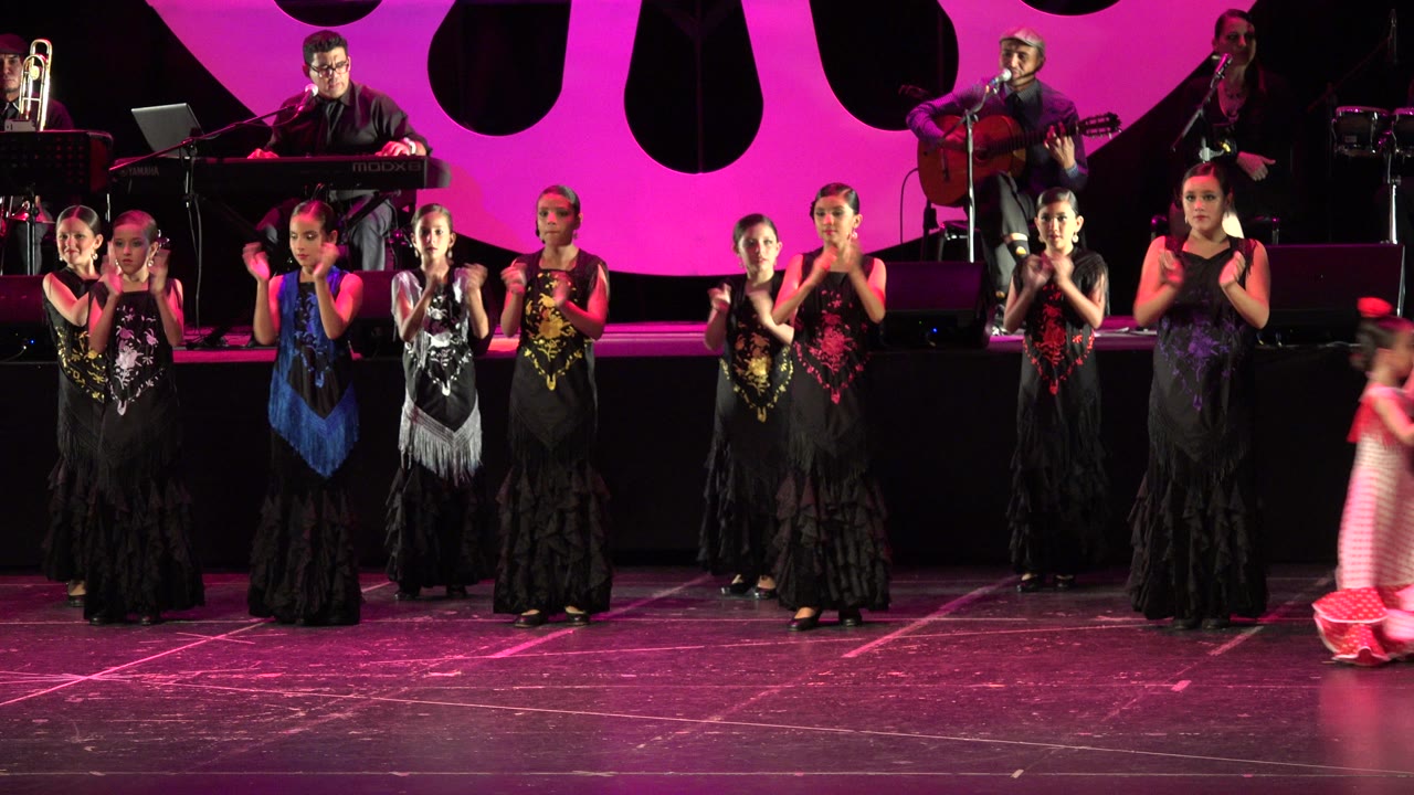 Young flamenco dancers clap on a pink-lit stage with a band behind them.