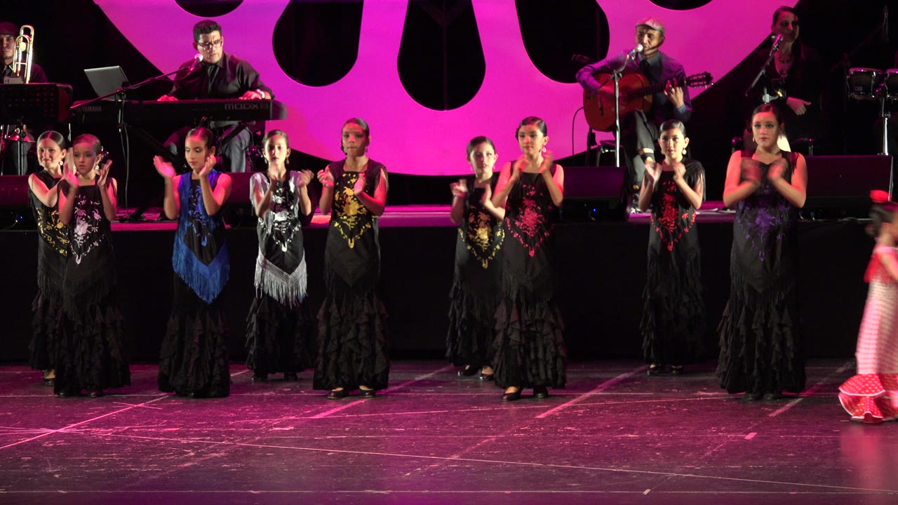 A younger child in a red and white outfit moves across the front of the Cabo San Lucas stage while the flamenco dance line continues clapping behind her.
