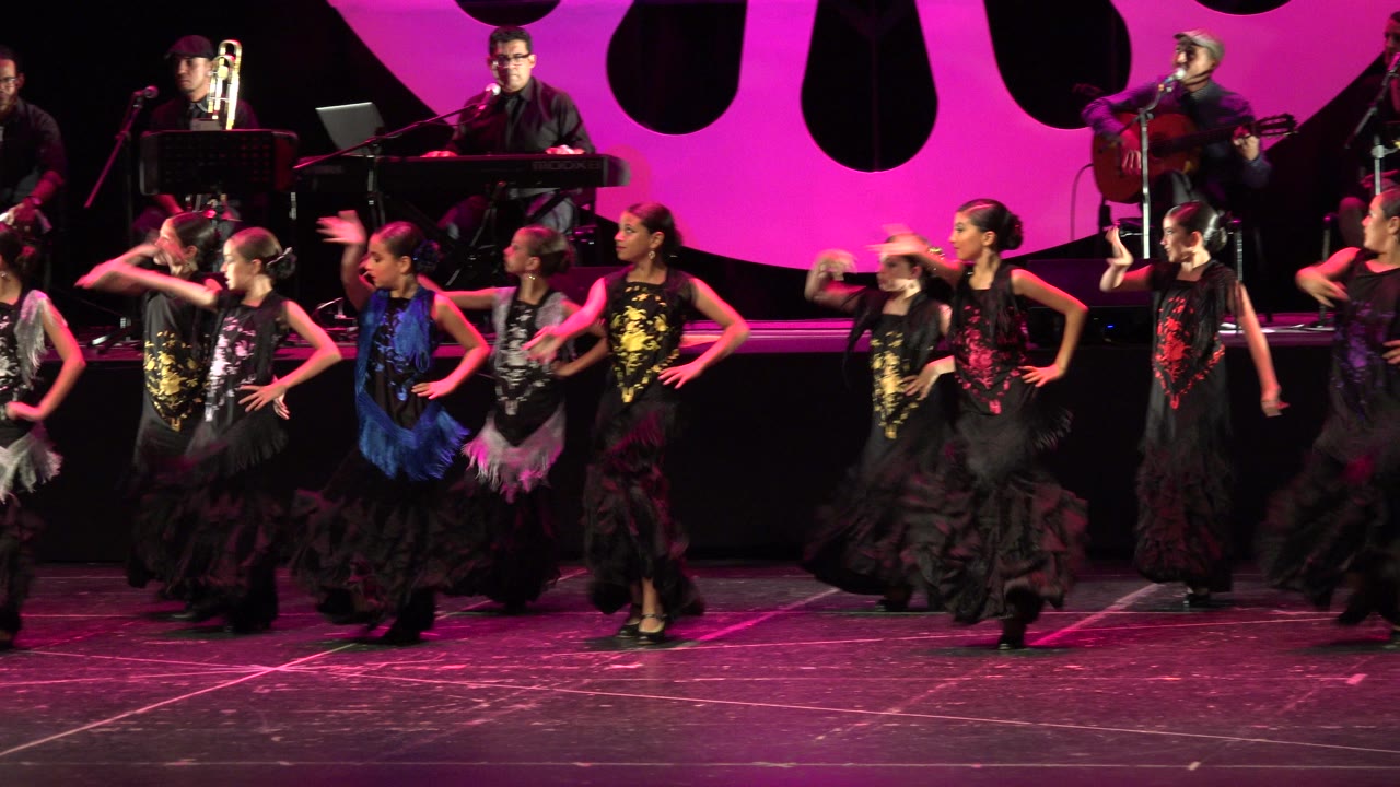 Children in colorful flamenco-style dresses dance on a magenta-lit stage with musicians behind them.