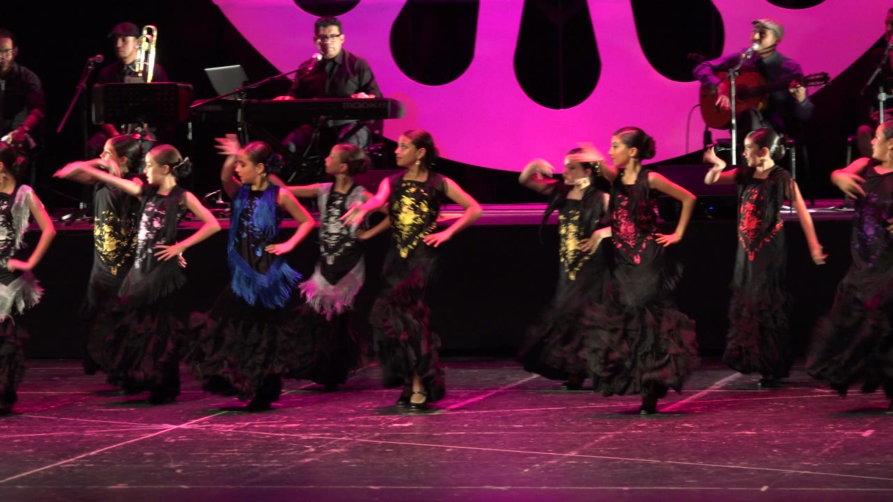 The dance group in Cabo San Lucas holds flamenco-style arm positions across the stage, with a trombone, keyboard, microphones, and magenta set pieces behind them.