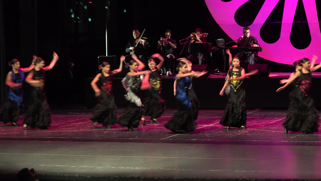 Girls in dark stage costumes dance in front of seated musicians.