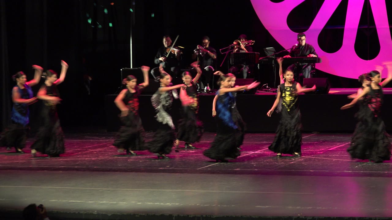 Young dancers in black dresses move across the Cabo San Lucas stage as the live musicians remain visible behind them and the magenta backdrop fills the right side.