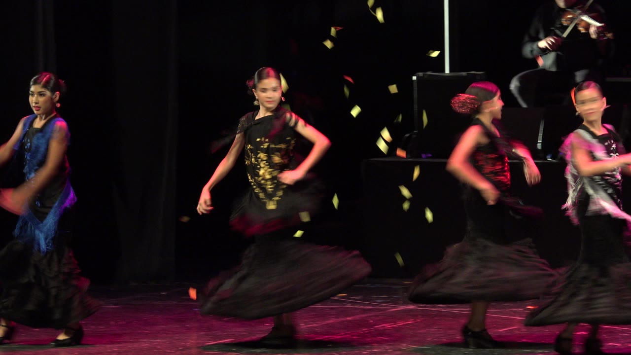 Children perform a choreographed dance on a dark stage in Cabo San Lucas, Mexico, with black ruffled skirts, colored costume panels, pink lighting, and falling confetti.