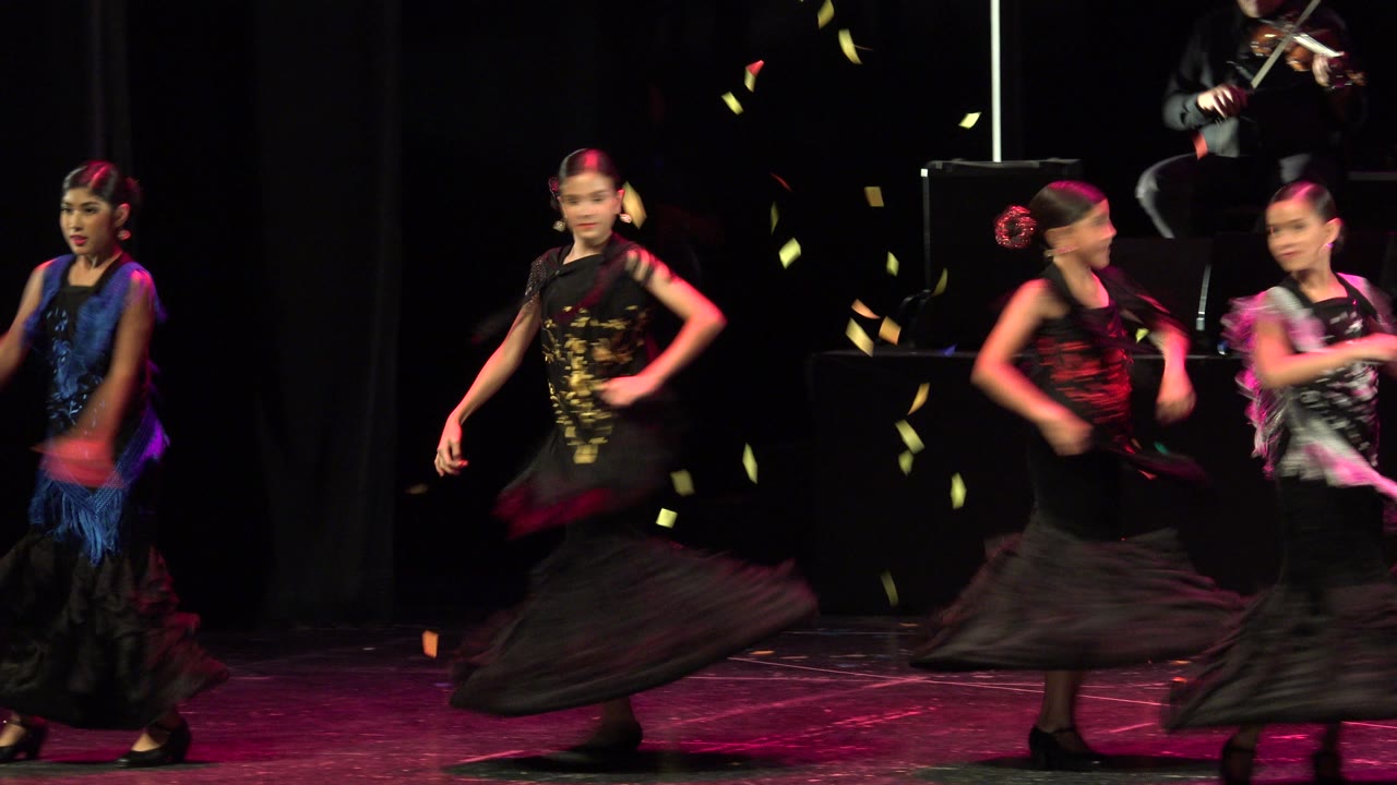 A group of child dancers crosses the stage in Cabo San Lucas, Mexico, while a seated violinist is partly visible on a raised platform behind them.