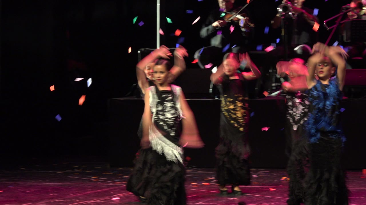 Child dancers move with raised arms as confetti falls on stage.