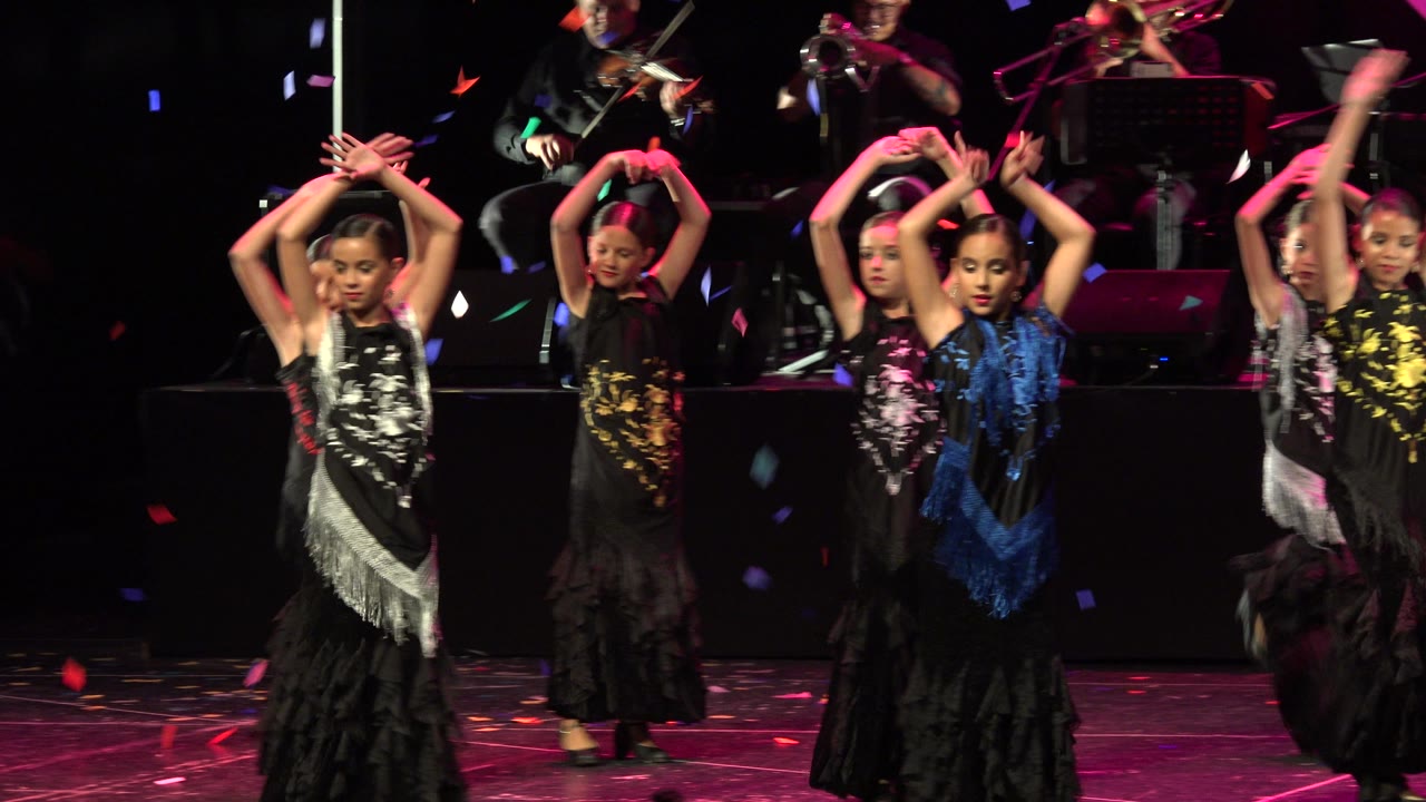 Two seconds in, the dancers hold overhead arm positions in a tighter formation on the Cabo San Lucas stage, with violin and brass players visible above the dark riser.