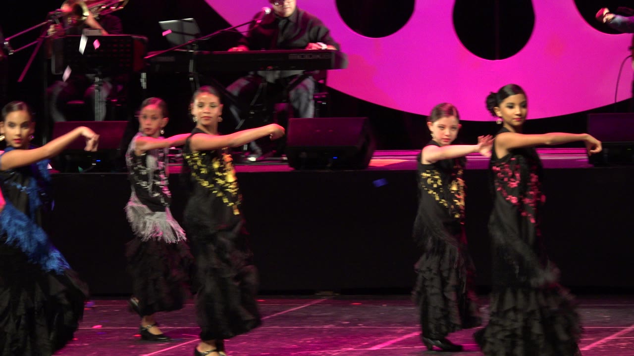 Young dancers in black dresses perform on a magenta-lit stage.