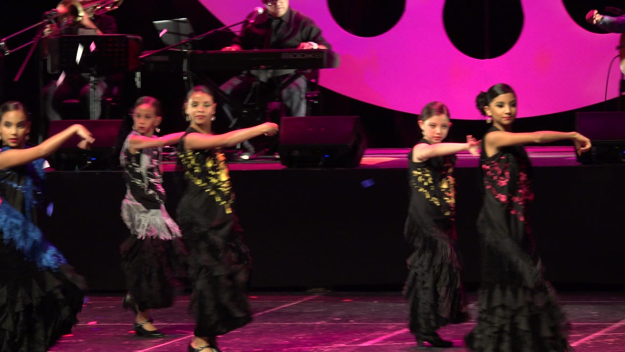 A line of young performers in black dresses continues a synchronized arm pose on the Cabo San Lucas stage, with a keyboard player and microphones in the background.