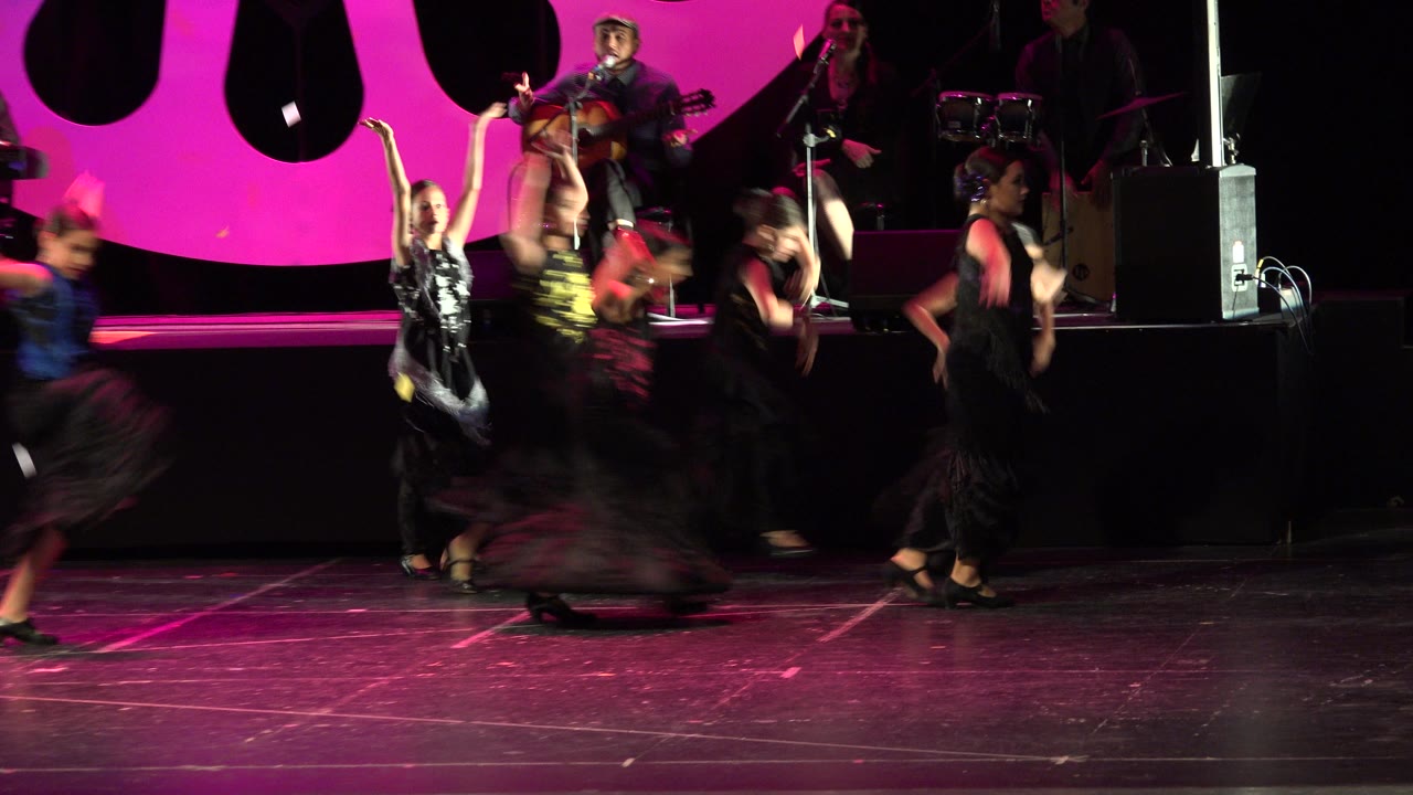 Fast-moving dancers pass through the foreground as musicians remain seated on the raised stage in Cabo San Lucas.