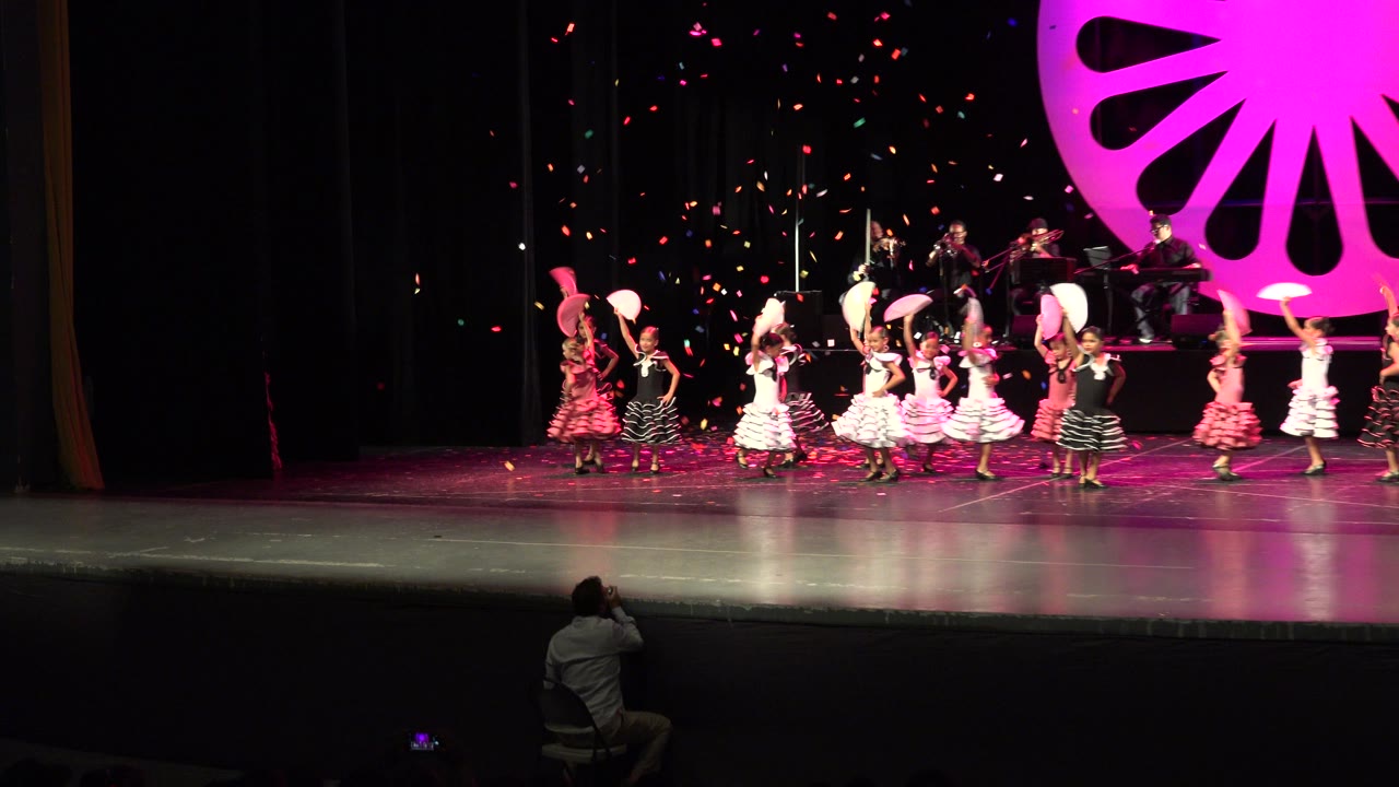 A group of children continues the Cabo San Lucas stage dance, with pink and white costumes clustered center stage and musicians visible on the rear platform.