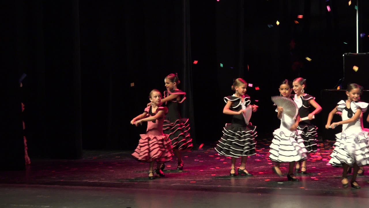 Girls in ruffled dresses dance on a dark stage with fans.