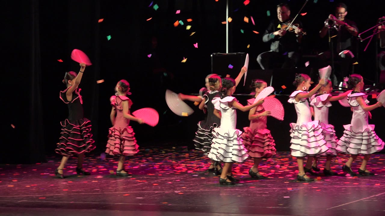 In Cabo San Lucas, young girls in ruffled dance costumes hold pink and white fans on a dark stage while confetti falls and musicians play behind them.