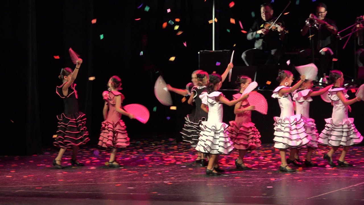A group of young dancers in pink, white, and black dresses cluster midstage in Cabo San Lucas, lifting fans while colorful confetti drops around them.
