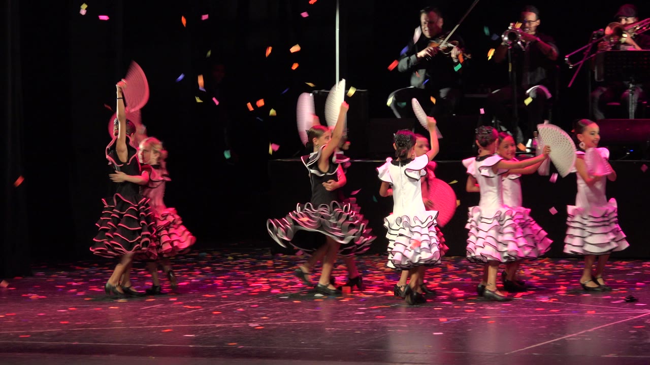 The dancers shift across the stage in Cabo San Lucas, with one black-and-pink skirt turning outward and several white fan props lifted near the musicians in back.