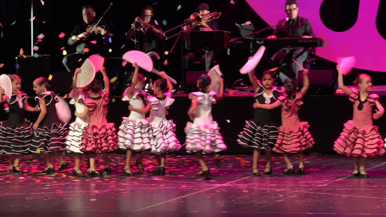 Children in red, white, pink, and black ruffled dresses perform with fans on a stage in Cabo San Lucas, Mexico, while musicians play on a raised platform behind them.