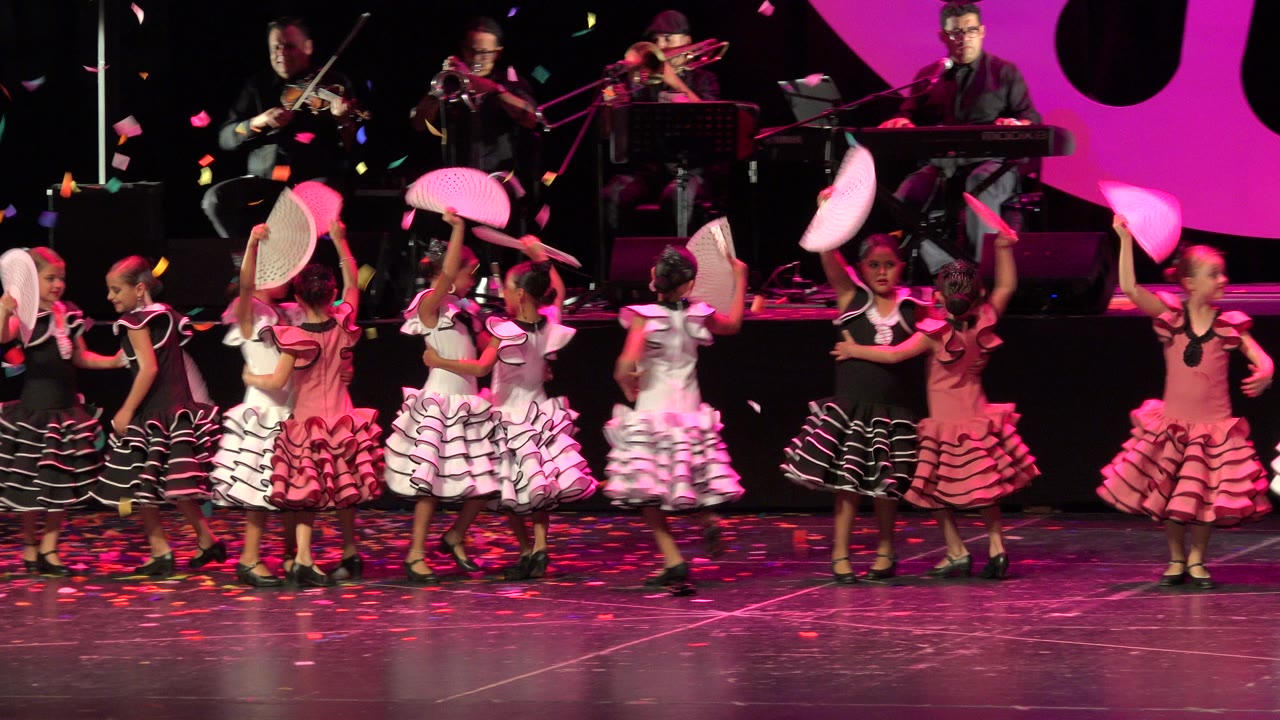 A line of young dancers in coordinated ruffled dresses raises fans during the Cabo San Lucas stage performance, with brass, violin, and keyboard players visible in the background.