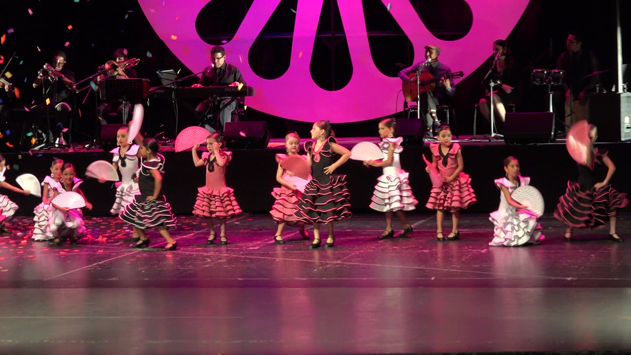 Children dancing on a theater stage with fans and musicians behind them.
