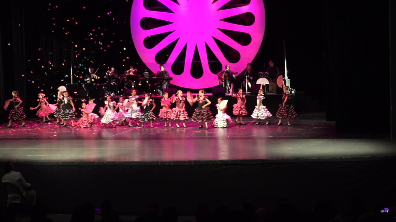 At the start of the Cabo San Lucas performance clip, children in colorful costumes spread across a stage while confetti falls in front of a large pink circular backdrop.