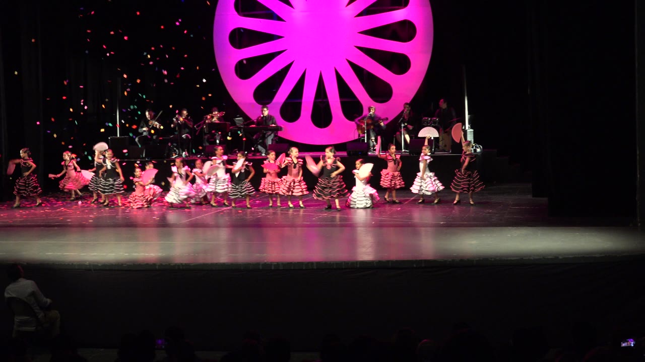 One second into the Cabo San Lucas clip, the children's dance group remains in a wide line while confetti drifts down and magenta stage light reflects on the floor.