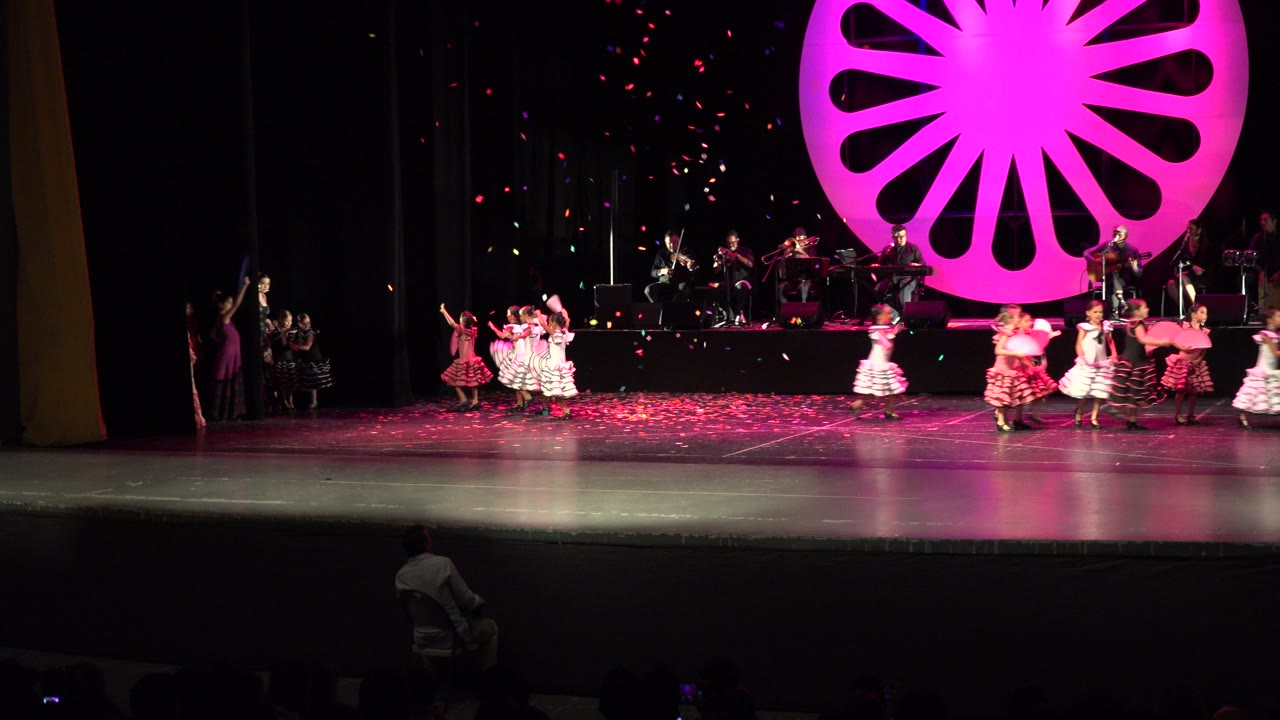 Wide theater stage with dancers, musicians, confetti, and a magenta backdrop.