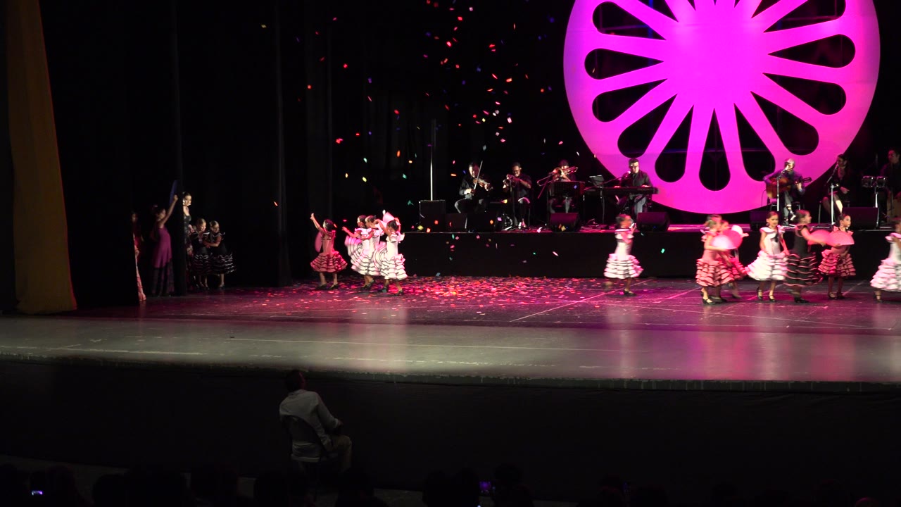 Women in red, yellow, green, and white dresses move across the Cabo San Lucas stage while confetti hangs in the dark background.