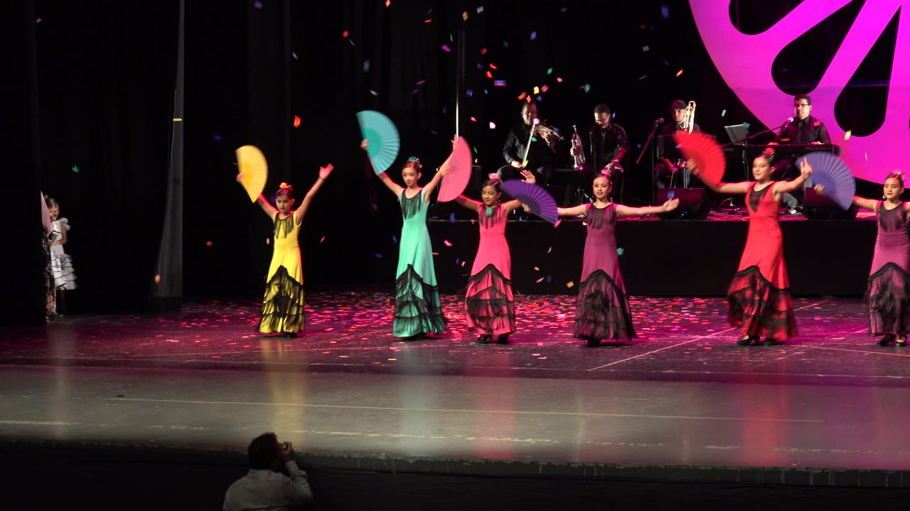 In Cabo San Lucas, Mexico, young girls in yellow, teal, pink, purple, and red dresses hold colored fans across a theater stage while musicians sit behind them.