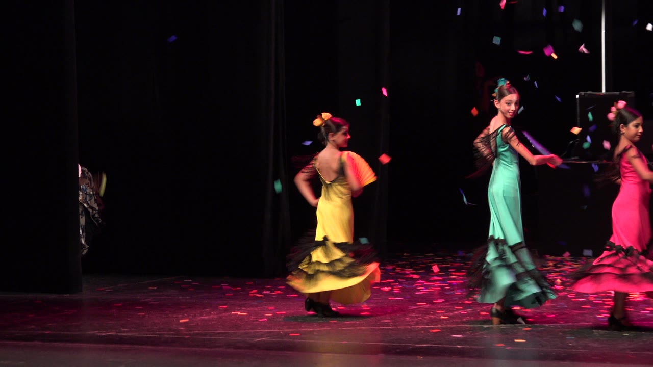 Three girls in colorful dresses dance on a dark stage with confetti overhead.