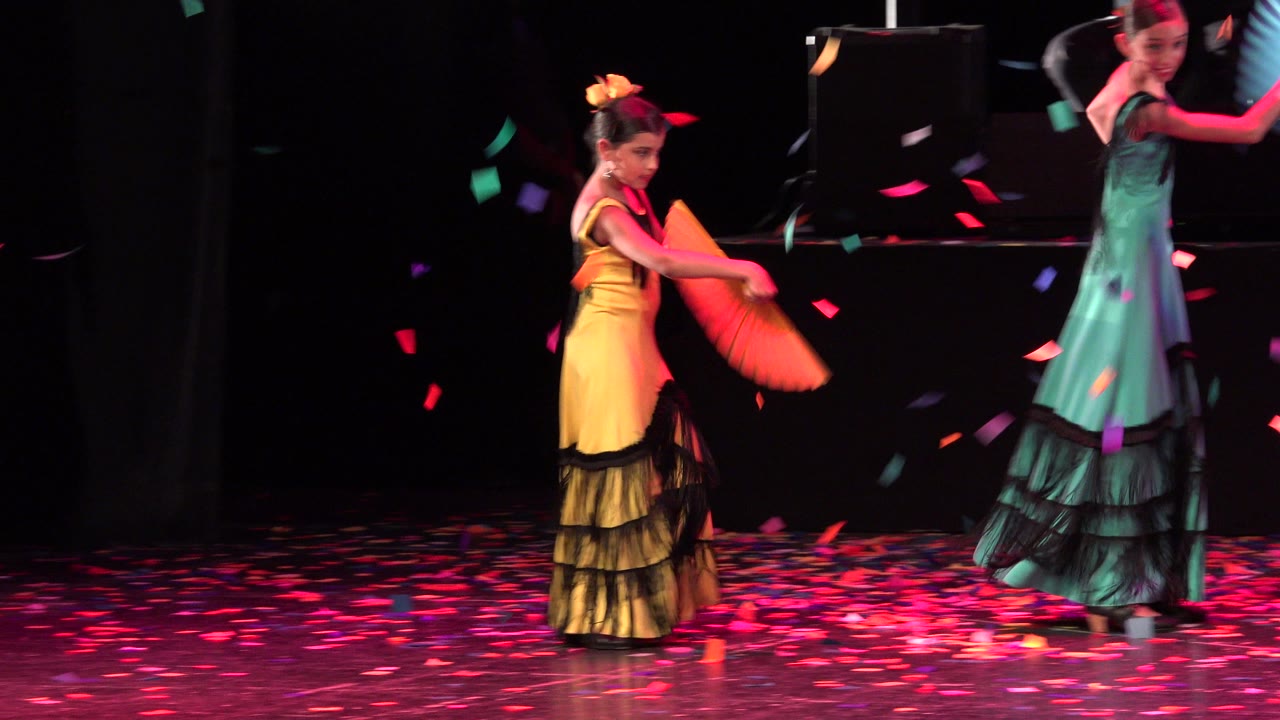Two young dancers in yellow and teal dresses perform with fans under falling confetti.