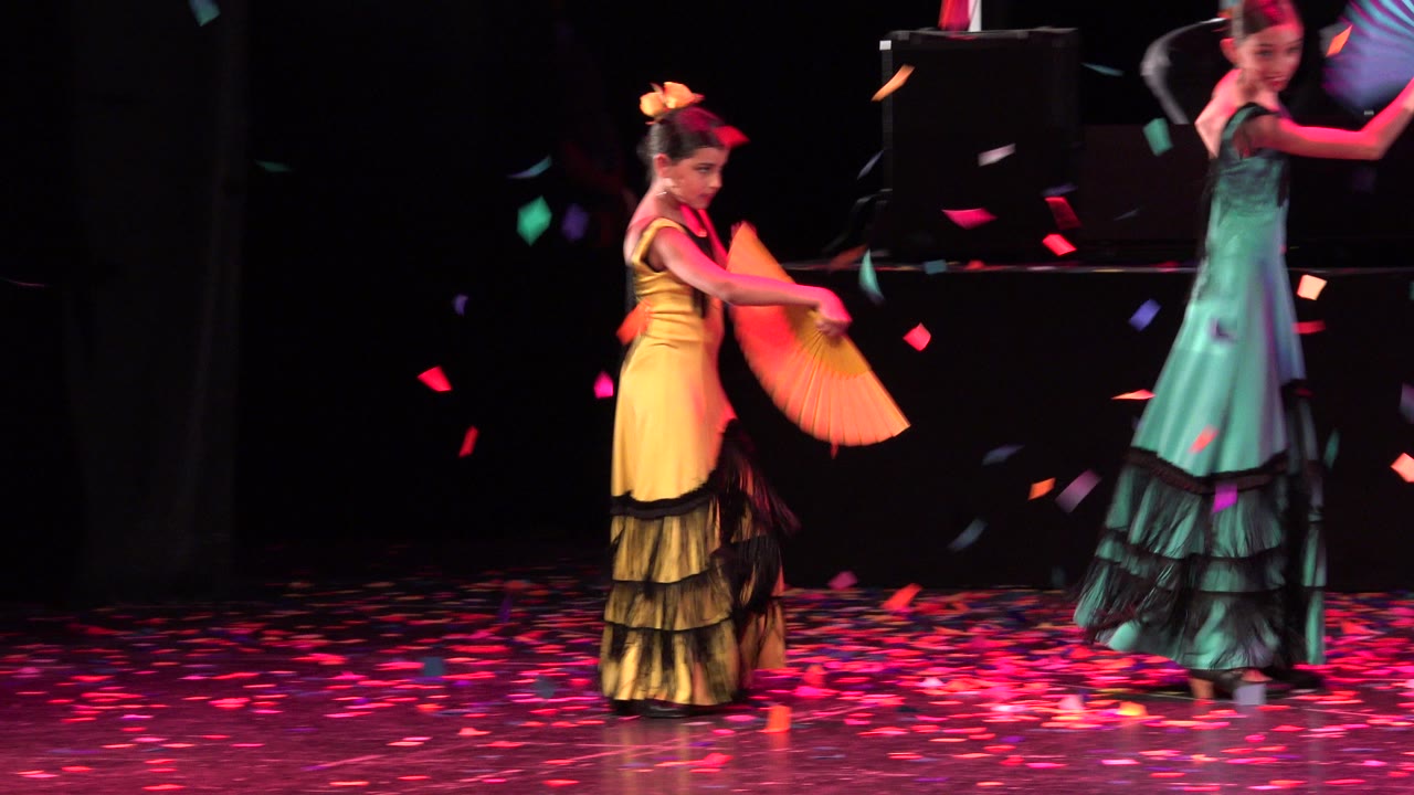 The yellow-dressed dancer lowers an orange fan while the teal-dressed dancer turns near the back of the Cabo San Lucas stage amid falling paper confetti.