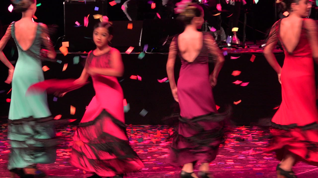 Girls in bright dresses dance on a stage with confetti falling.