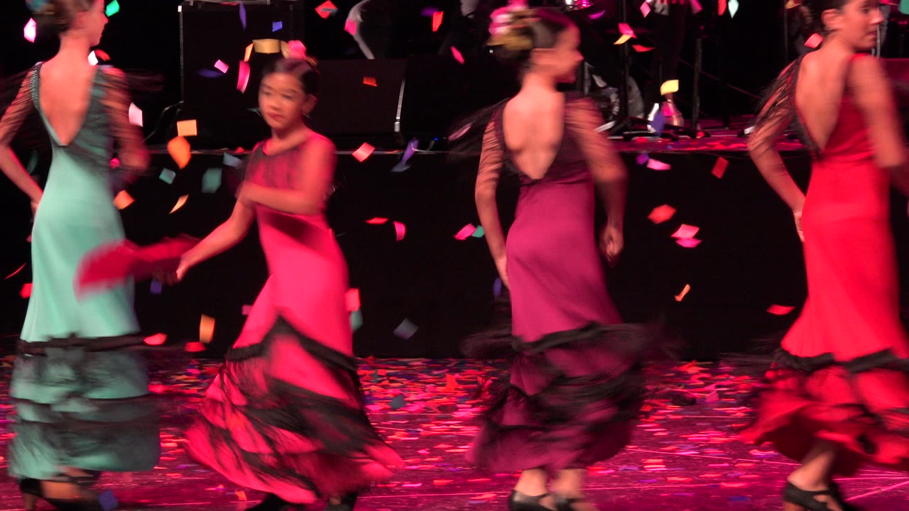 Children in red, purple, and teal ruffled dance dresses move across the stage in Cabo San Lucas, Mexico, with confetti visible in the air and on the floor.