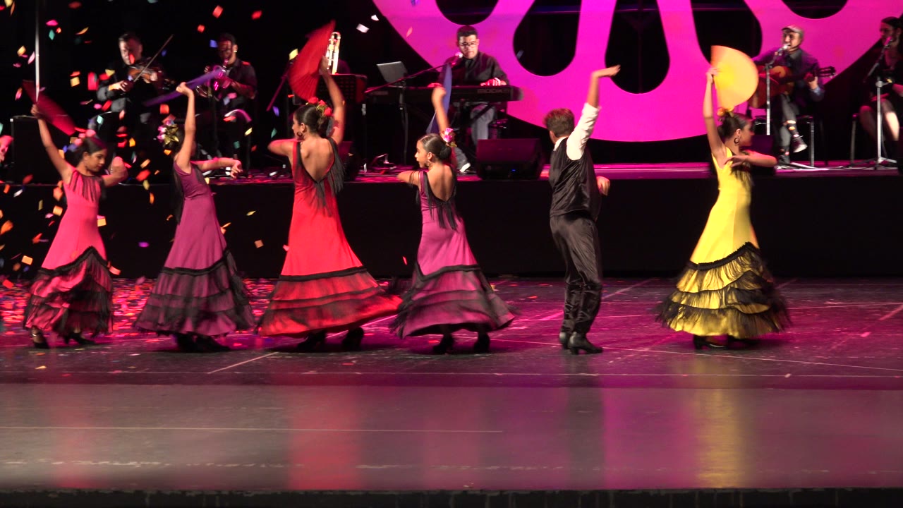 Young dancers twirl in colorful dresses on a stage with musicians behind them.