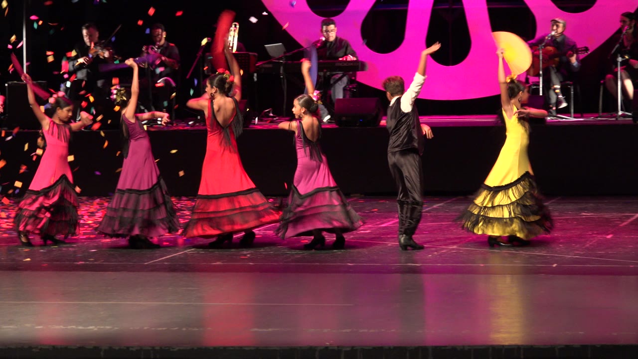 In Cabo San Lucas, Mexico, the dancers continue in a wide stage formation with raised arms, falling confetti, and seated musicians visible across the back.