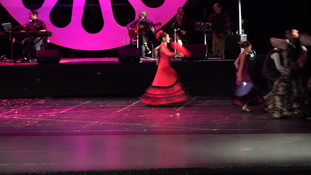 A dancer in a red dress crosses the Cabo San Lucas stage as other performers in black floral dresses blur past under pink stage light.