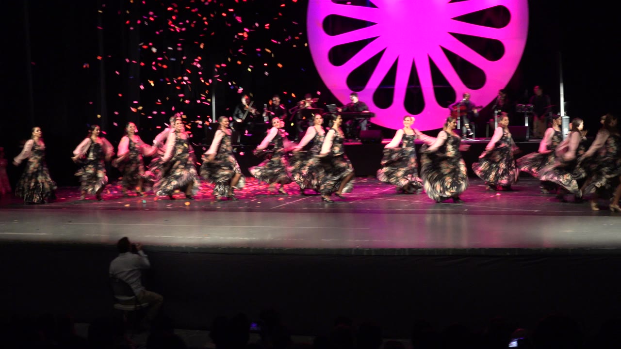 Wide stage view of women dancing under pink light with confetti behind them.