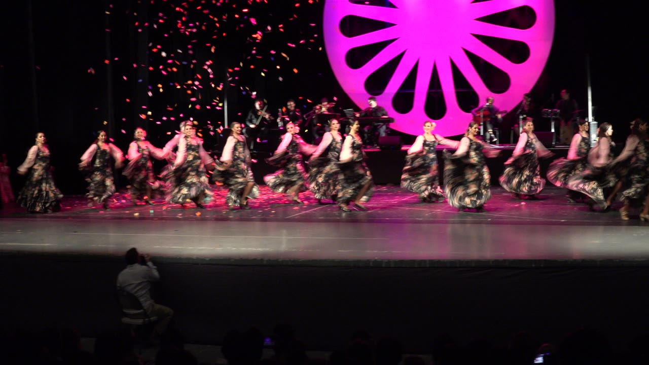 In Cabo San Lucas, Mexico, the dance line shifts across the stage with skirts in motion, musicians seated behind, and pink confetti visible against the dark background.