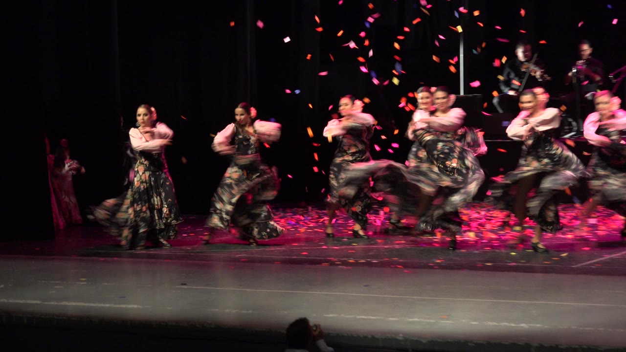 Dancers in floral dresses perform on a stage as confetti falls.