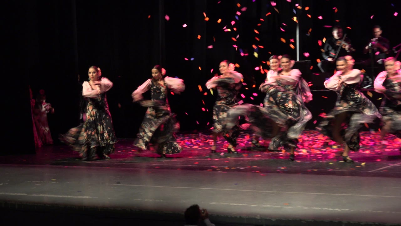 The Cabo San Lucas stage performance continues with dancers turning in floral dresses while pink and orange confetti hangs above the dark backdrop.