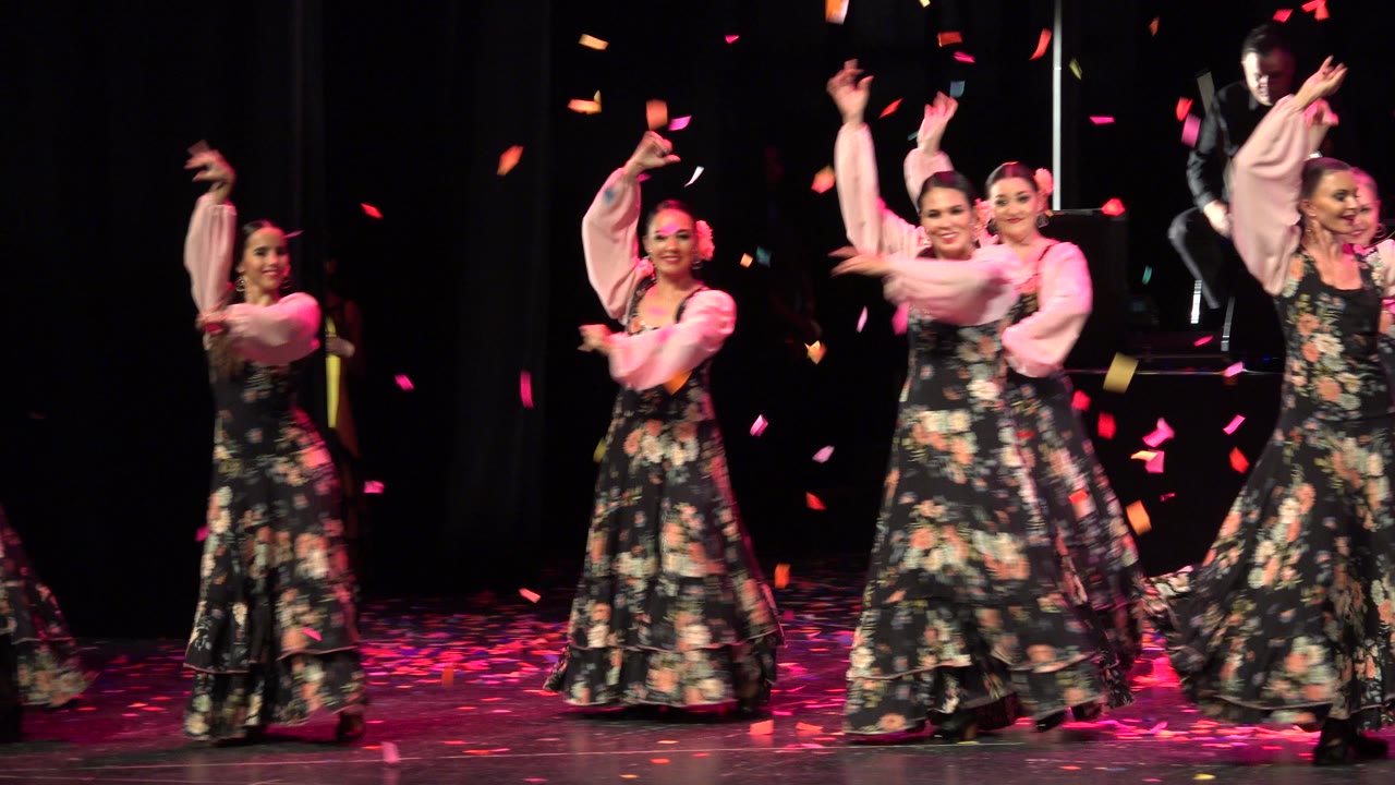 Women in floral dresses dance on a dark stage as confetti falls.