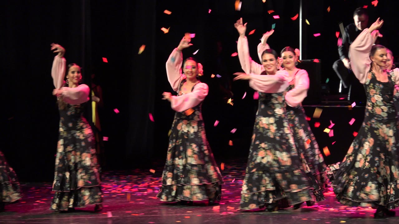 At 0:01, several visible dancers hold raised arm poses on the Cabo San Lucas stage as pink and orange confetti crosses the foreground.