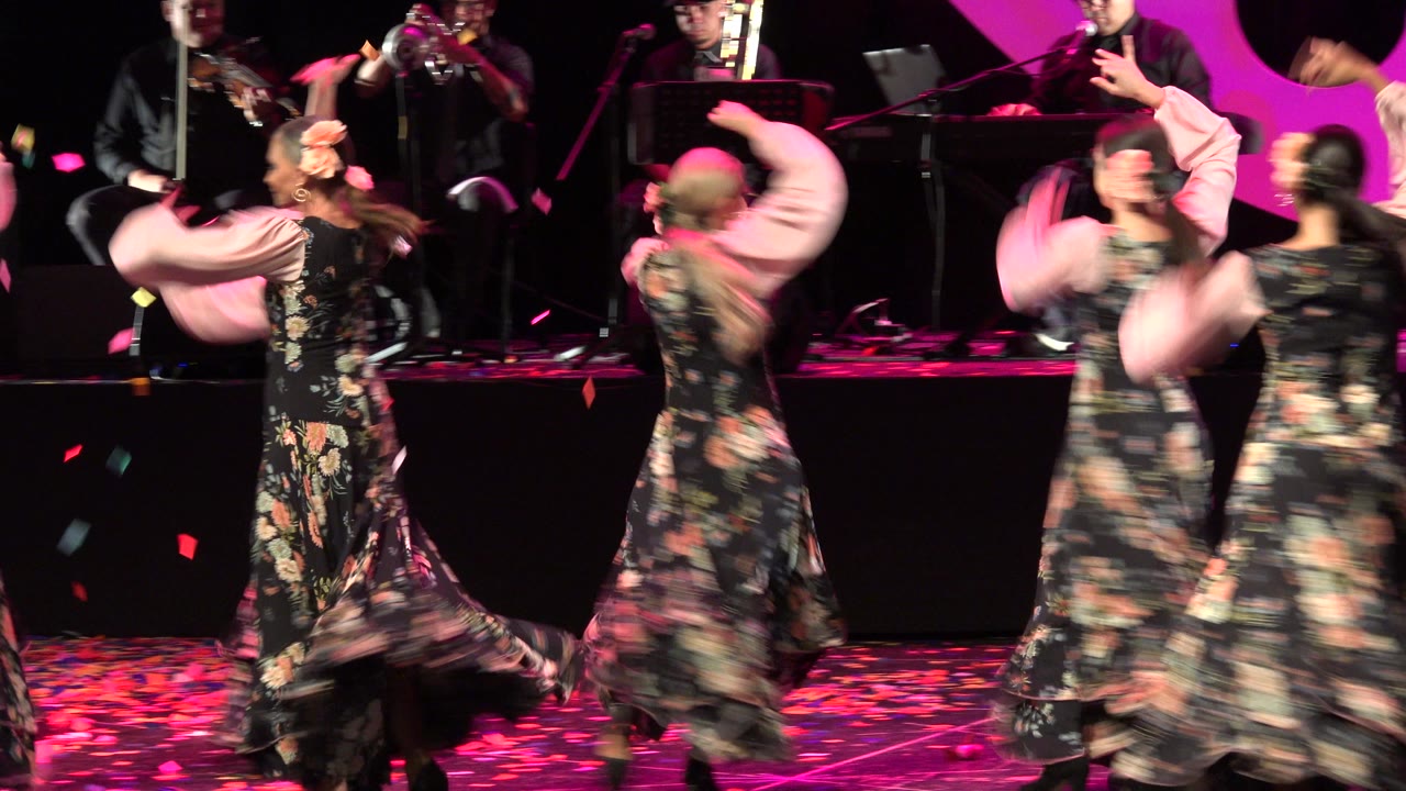 Flamenco dancers in floral dresses perform on a magenta-lit stage with musicians behind them.