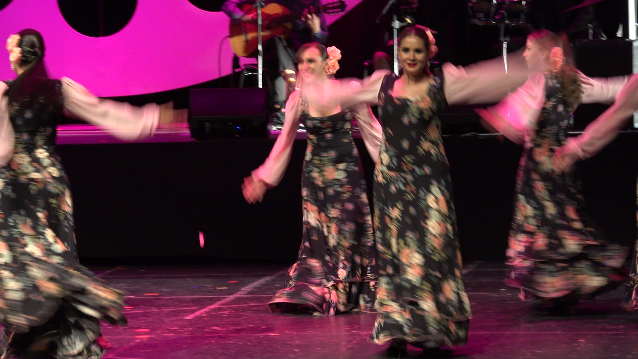 Women in long black floral dresses dance on a stage in Cabo San Lucas, Mexico, with pink lighting and musicians partly visible behind them.