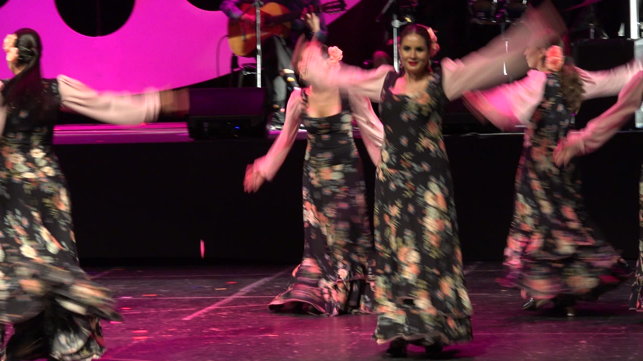 The stage dance continues in Cabo San Lucas, Mexico, with several women in floral dresses lifting their arms while performers and instruments remain in the background.