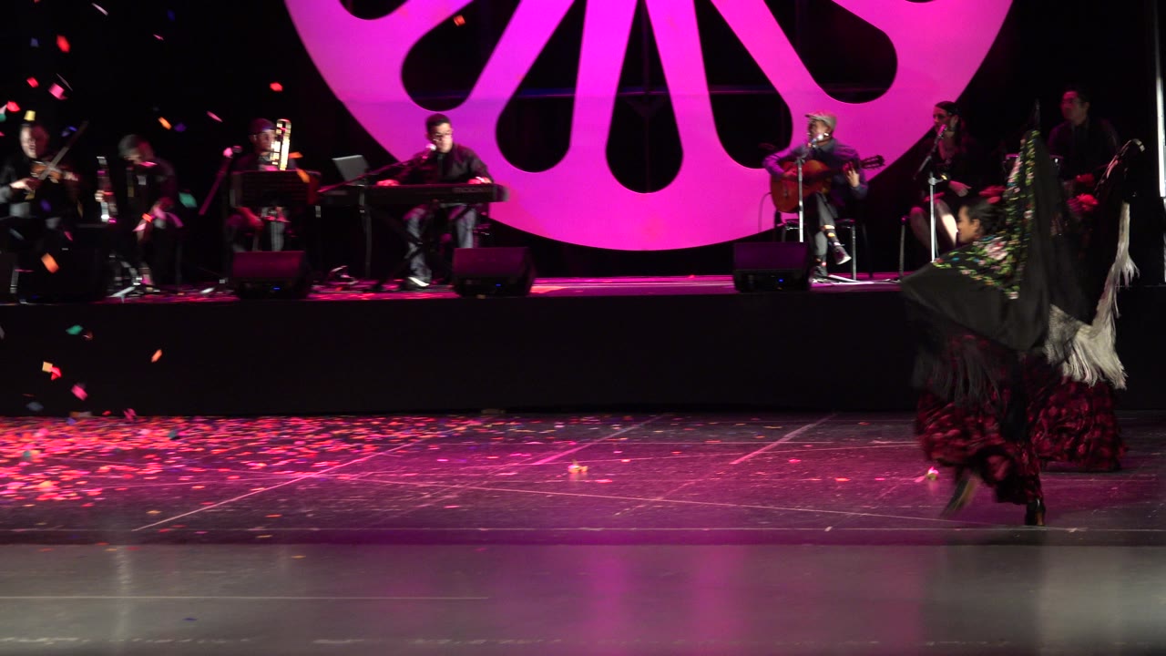 A dancer in a dark shawl and red skirt shifts toward the right side of the Cabo San Lucas stage while the seated band and pink circular backdrop remain in the background.