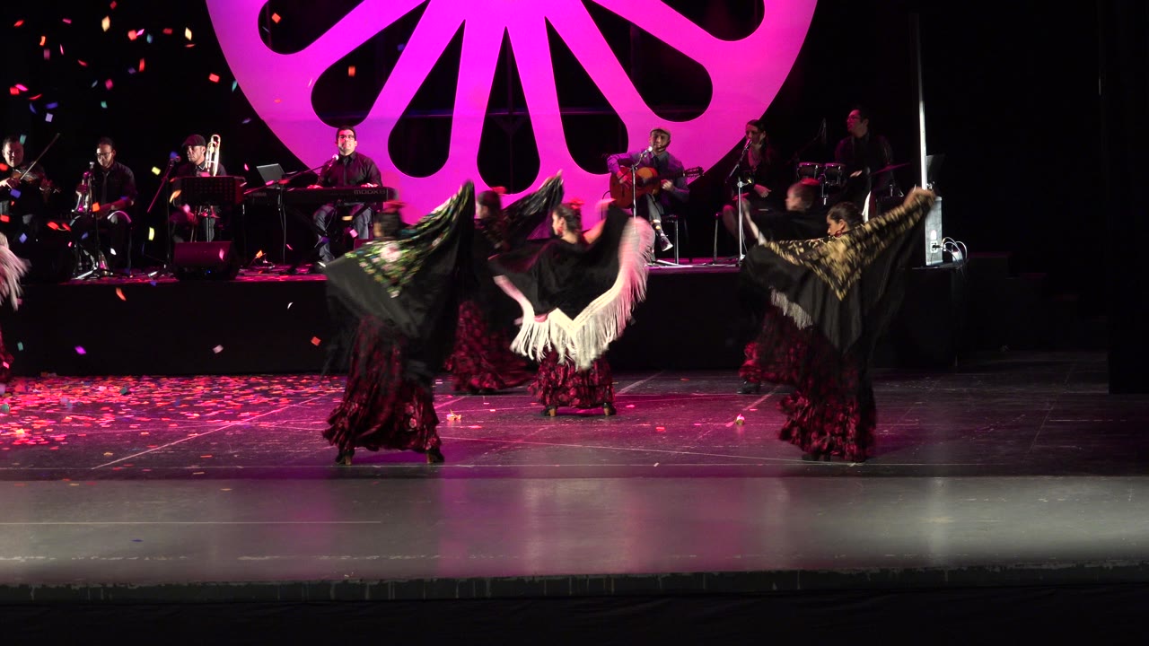 In Cabo San Lucas, Mexico, dancers in dark red skirts and black fringed shawls perform across a stage while musicians sit and stand behind them under a large magenta shell-shaped backdrop.