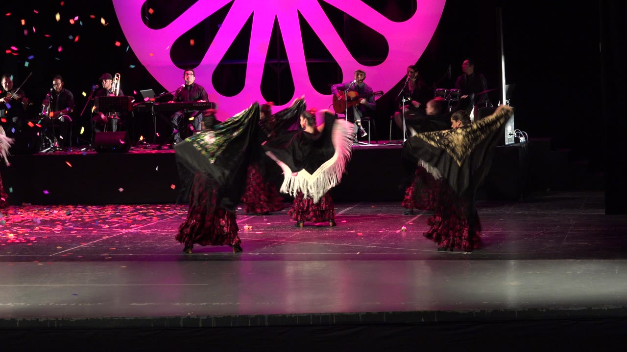 A group of dancers continues the Cabo San Lucas stage performance, with white and black shawl fringe lifted in motion and band members framed beneath the bright magenta backdrop.