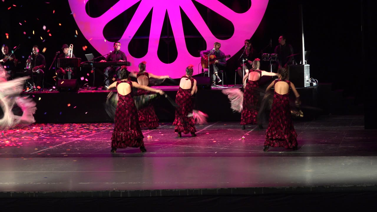 Dancers turn with their backs partly visible on a magenta-lit stage.