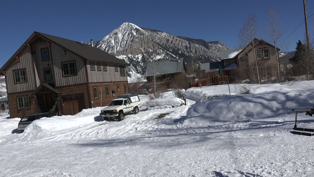 Opening frame of a Crested Butte, Colorado snowy residential street with a wood-sided house at left, a white parked car near the snowbank, mountain ridge behind, and no visible people.